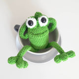 Happy loking green amigurumi crochet frog sitting in a cup and saucer looking up at the viewer