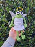 Hand holding a green crochet angel frog toy against a leafy background