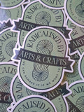Stack of 'Radicalised by Arts & Crafts' stickers on a green background