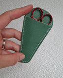 A hand holding a small green leather sheath containing red-handled scissors against a textured white wall.