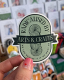 Sticker with 'Radicalised by Arts & Crafts' text held by a hand in front of a blurred store background.